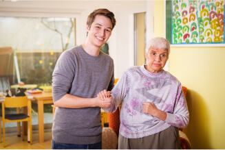 Ein junger Mann mit kurzen braunen Haaren und einem grauen Pullover hält die Hand einer älteren Frau mit kurzen weißen Haaren. Sie trägt einen lilafarben gemusterten Pullover und eine graue Hose. Beide lächeln freundlich. Sie befinden sich in einem gemütlichen Raum mit Holzmöbeln und bunten Bildern an der Wand. Die Atmosphäre wirkt warm und einladend.