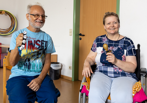 Ein Mann und eine Frau in Rollstuhl nehmen an einer Musikstunde teil. Er hält eine Schelle und sie eine Rassel in der Hand. Sie sehen zufrieden und glücklich aus.