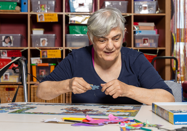 Eine Frau ist mit einem Puzzle beschäftigt. Sie sitzt an einem Tisch in der Tagesstruktur, hat kurze graue Haare und ein blaues Oberteil.