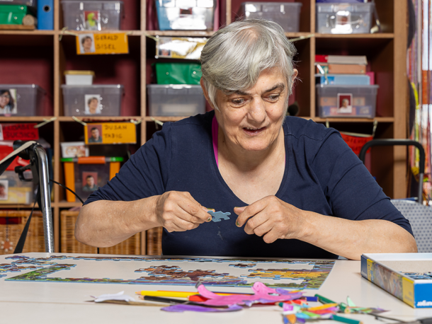 Eine Frau ist mit einem Puzzle beschäftigt. Sie sitzt an einem Tisch in der Tagesstruktur, hat kurze graue Haare und ein blaues Oberteil.