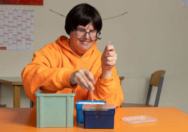 Eine Frau im orangenen Pullover und mit Brille erledigt eine Auftragsarbeit. Sie schlichtet Spitzen für ein Labor in einen Behälter.