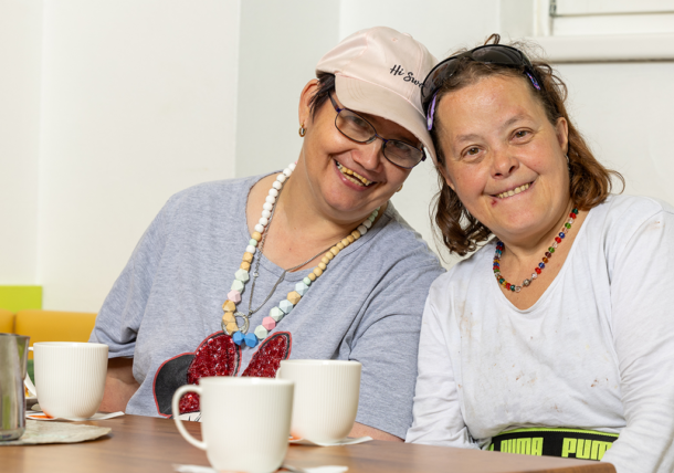 Zwei Frauen sitzen zufrieden beim Kaffee zusammen. Sie tragen Ketten um den Hals, eine hat eine Kappe auf und lächeln in die Kamera. Vor ihnen auf einem Tisch stehen Kaffeetassen und ein Milchkännchen.
