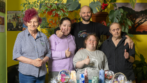 Eine Gruppe KundInnen steht zu hinter einem Tisch zusammen und alle schauen freundlich in die Kamera. Am Tisch vor ihnen sind die handgefertigten Seifen und Kerzen aufgebaut, die in der Tagesstruktur hergestellt werden.