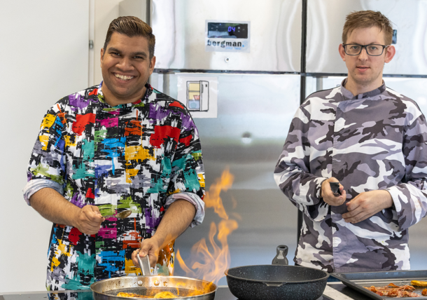 Zwei Männer in bunt gemusterten Kochjacken arbeiten in einer Kueche der Lebenshilfe Wien. Sie braten Fleisch an. Vor ihnen auf einer Kochplatte stehen zwei Pfannen, aus einer davon steigen Flammen empor.
