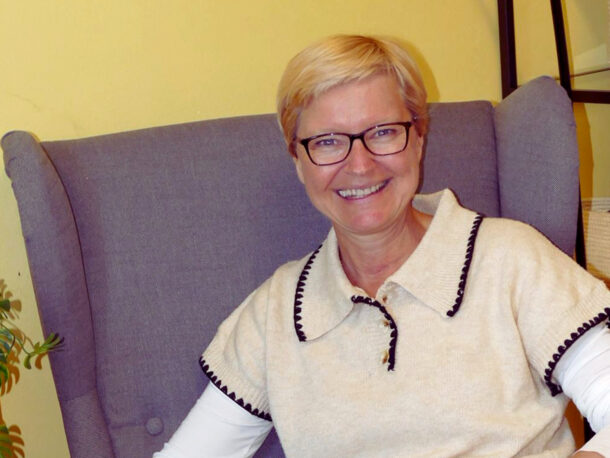Eine lächelnde Person mit kurzen blonden Haaren und Brille sitzt in einem grauen Sessel vor einer gelben Wand, bekleidet mit einem von Kaltenbrunner inspirierten cremefarbenen Oberteil mit schwarzem Saum und einem weißen langärmeligen Hemd. In der Nähe sind eine Pflanze und geflochtene Körbe zu sehen.
