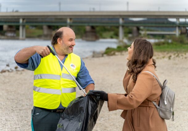 Zwei Personen sammeln gemeinsam Müll auf der Donauinsel. Der Mann ist ein Kunde der Lebenshilfe Wien und die Frau ist eine Mitarbeiterin einer Firma. Der Mann hält eine Zange, die Frau einen Müllsack. Sie haben Spaß an der gemeinsamen Aktion und sind in ein Gespräch vertieft.