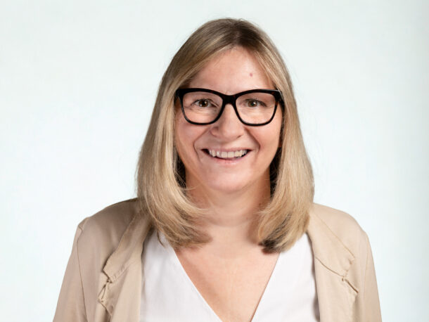 Eine Frau mittleren Alters mit schulterllangen blonden Haaren und dunkler Brille lächelt freundlichlich in die Kamera