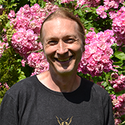 Der Mitarbeiter Robert Jez schaut in die Kamera. Er hat zusammengebundene Haare und steht vor einem blühenden Strauch. Er hat ein schwarzes T-Shirt mit einem Bienen-Aufdruck an.