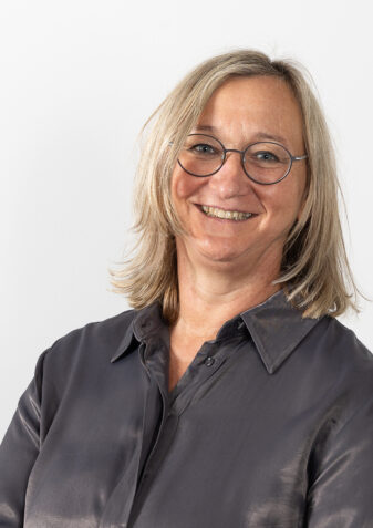 Eine Frau mittleren Alters lächelt in die Kamera. Sie hat längere, blone Haare und trägt eine Brille und eine dunkle Bluse