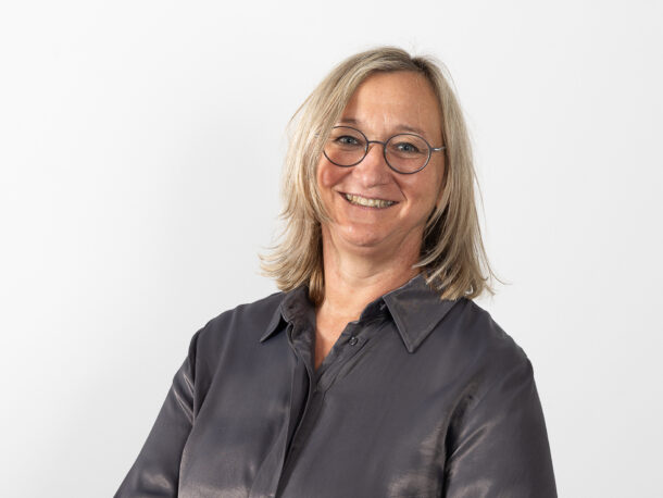 Eine Frau mittleren Alters lächelt in die Kamera. Sie hat längere, blone Haare und trägt eine Brille und eine dunkle Bluse