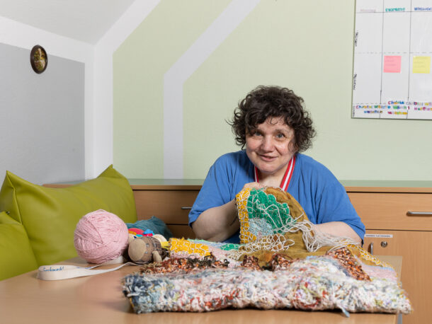 Eine lächelnde Frau mit blauem T-Shirt sitzt an einem Tisch zeigt ihr buntes Handarbeits-Projekt in die Kamera. Auf der Bank neben ihr befinden sich zwei grüne Pölster und auf der Wand hinter ihr hängt ein Kalender.