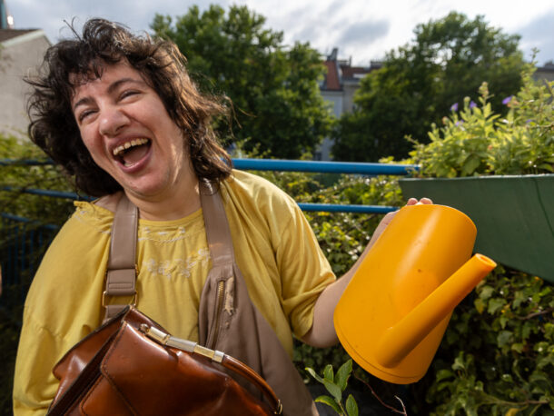 Eine lachende Frau steht auf einer begrünten Terrasse und hält eine gelbe Gießkanne in der Hand. Sie hat braune Haare und trägt ein gelbes T-Shirt.