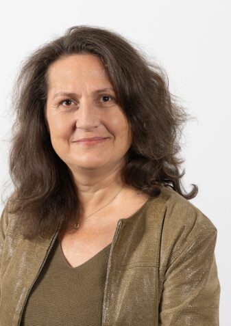 Eine dunkelhaarige Frau mit olivfarbenen Oberteil sieht freundlich in die Kamera