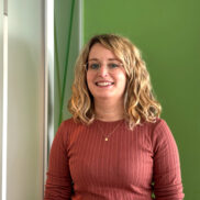 Porträt einer lächelnden Frau mit blonden, welligen Haaren, die eine Brille trägt. Sie hat ein rostfarbenes, geripptes Oberteil an und trägt eine filigrane Halskette. Der Hintergrund ist in hellgrünen und weißen Farbtönen gestaltet.