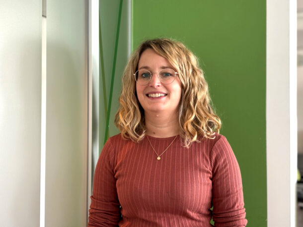 Porträt einer lächelnden Frau mit blonden, welligen Haaren, die eine Brille trägt. Sie hat ein rostfarbenes, geripptes Oberteil an und trägt eine filigrane Halskette. Der Hintergrund ist in hellgrünen und weißen Farbtönen gestaltet.
