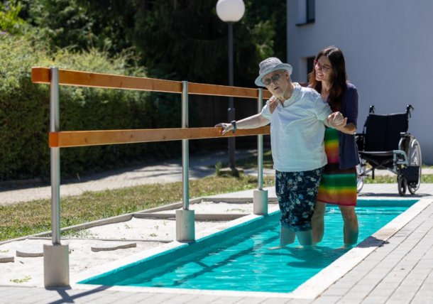 Zwei Frauen kneippen im Garten einer Wohngemeinschaft der Lebenshilfe Wien. Die ältere Dame mit Hut wird von einer Mitarbeiterin beim Gehen unterstuetzt.