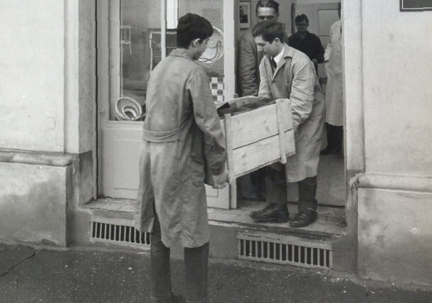 Eine alte Schwarzweiß Aufnahme aus dem Jahr 1966 zeigt den Eingang der ersten Werkstatt im 16. Wiener Gemeindebezirk. Zwei Männer transportieren ein Holzmöbelstück durch die Eingangstür.
