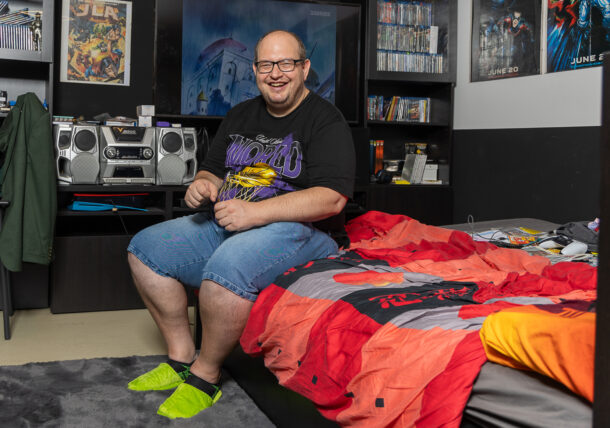 Ein Mann mit kurzen Haaren und Brille sitzt auf seinem Bett. Im Hintergrund sieht man eine Stereobox, eine DVD-Sammlung und viele Film-Poster.
