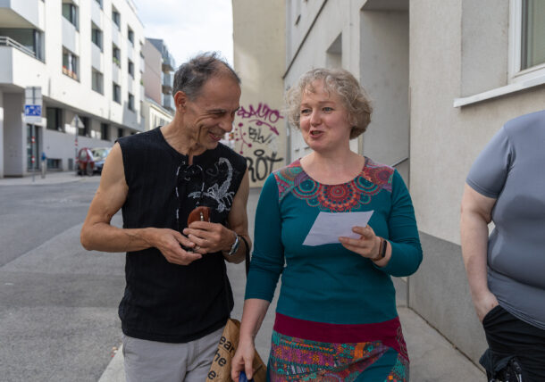 Ein Mann und eine Frau gehen am Gehsteig, sie unterhalten sich. Die Frau hat einen Zettel in der Hand, der Mann einen Schlüsselbund.