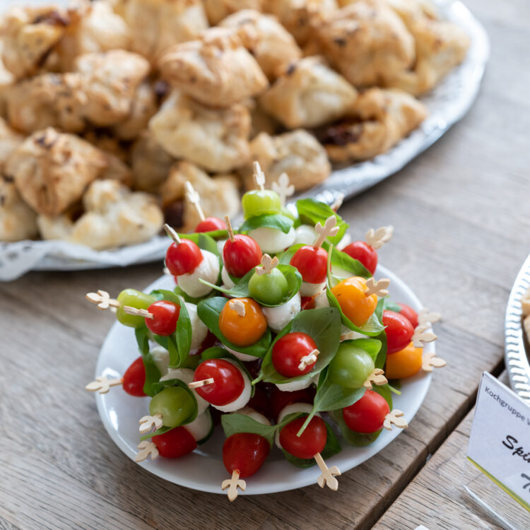 Das Foto zeigt kalte Häppchen, pikantes Fingerfood, dass die Kochgruppe der Lebenshilfe Wien zubereitet hat. Man sieht Spieße mit Mozzarella und Tomaten sowie gefüllte Blätterteigtaschen auf einen Tisch stehen.