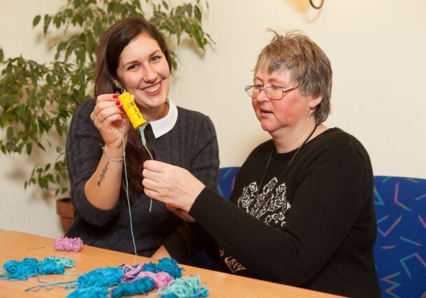 Eine freiwillige Mitarbeiterin (links im Bild) hält eine Strickliesel in der Hand. Neben ihr sitzt eine Kundin aus der Lebenshilfe Wien, die sich über das gemeinsame Handarbeiten freut.