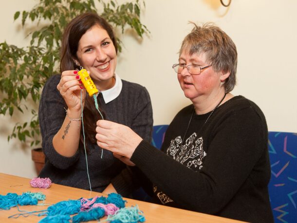 Eine freiwillige Mitarbeiterin (links im Bild) hält eine Strickliesel in der Hand. Neben ihr sitzt eine Kundin aus der Lebenshilfe Wien, die sich über das gemeinsame Handarbeiten freut.