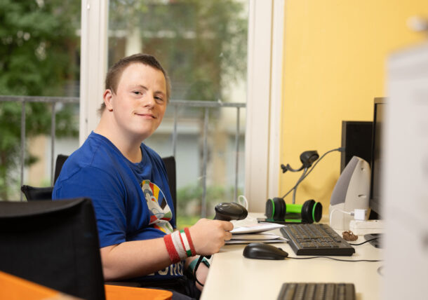 Ein junger Mann mit Down-Syndrom sitzt an einem Schreibtisch in einem Büro. Er trägt ein blaues T-Shirt mit einem bunten Logo und mehrere Armbänder. Er hält einen Barcodescanner in der Hand und lächelt freundlich in die Kamera. Auf dem Schreibtisch befinden sich eine Tastatur, eine Maus, ein Monitor sowie weitere Bürogegenstände. Im Hintergrund ist ein großes Fenster mit Blick auf eine grüne Umgebung zu sehen.