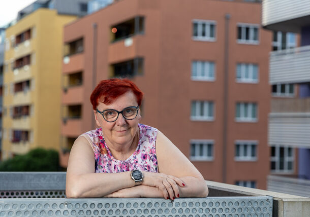 Eine ältere Frau mit kurzen roten Haaren und Brille lehnt sich auf das Geländer eines Balkons. Sie trägt ein ärmelloses, bunt gemustertes Oberteil und eine Armbanduhr. Im Hintergrund sind mehrstöckige Wohngebäude in verschiedenen Farben zu sehen. Sie blickt freundlich in die Kamera.