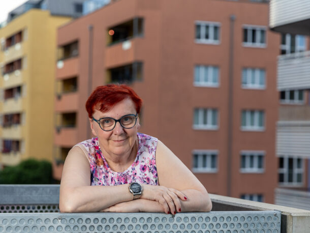 Eine ältere Frau mit kurzen roten Haaren und Brille lehnt sich auf das Geländer eines Balkons. Sie trägt ein ärmelloses, bunt gemustertes Oberteil und eine Armbanduhr. Im Hintergrund sind mehrstöckige Wohngebäude in verschiedenen Farben zu sehen. Sie blickt freundlich in die Kamera.