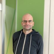 Der Mitarbeiter Matthias Papst schaut freundlich in die Kamera, er ist ein Mann mittlern Alterns mit Glatze, Brille und Dreitagesbart. Er traegt einen dunklen Sweater und steht vor einer grünen Wand.