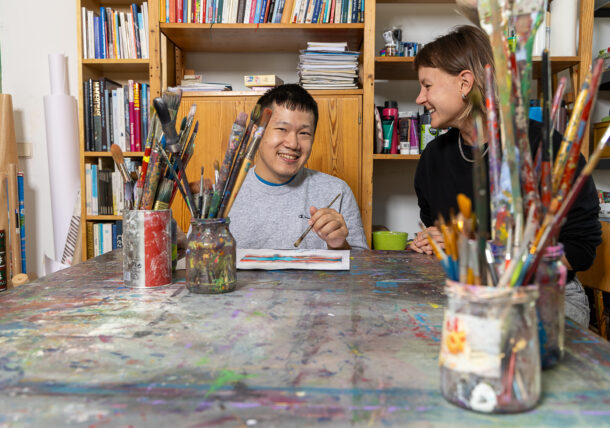 Das Foto zeigt zwei Personen an einem Tisch sitzend in einer Tagesstruktur der Lebenshilfe. Der junge Mann, links am Bild, ist Kunde der Lebenshilfe Wien und malt im Atelier. Er wird von einer Praktikantin unterstuetzt bei seiner kreativen Arbeit mit Pinsel und Farben.