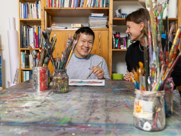 Das Foto zeigt zwei Personen an einem Tisch sitzend in einer Tagesstruktur der Lebenshilfe. Der junge Mann, links am Bild, ist Kunde der Lebenshilfe Wien und malt im Atelier. Er wird von einer Praktikantin unterstuetzt bei seiner kreativen Arbeit mit Pinsel und Farben.