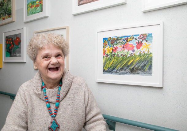 Einen ältere Fraue lächelt freundlich, sie steht vor einem gemalten Bild mit Blumen an einer weißen Wand.
