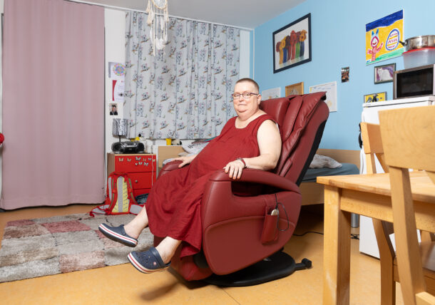 Auf dem Bild sitzt eine Frau mit kurzen Haaren, Brille und einem roten Kleid auf einem roten Ledersessel in ihrem Zimmer. Das Zimmer ist bunt und individuell eingerichtet. An einer blauen Wand im Hintergrund hängen mehrere Bilder, die der Szene eine persönliche und lebendige Atmosphäre verleihen.