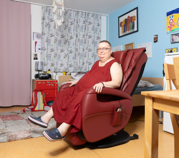 Auf dem Bild sitzt eine Frau mit kurzen Haaren, Brille und einem roten Kleid auf einem roten Ledersessel in ihrem Zimmer. Das Zimmer ist bunt und individuell eingerichtet. An einer blauen Wand im Hintergrund hängen mehrere Bilder, die der Szene eine persönliche und lebendige Atmosphäre verleihen.