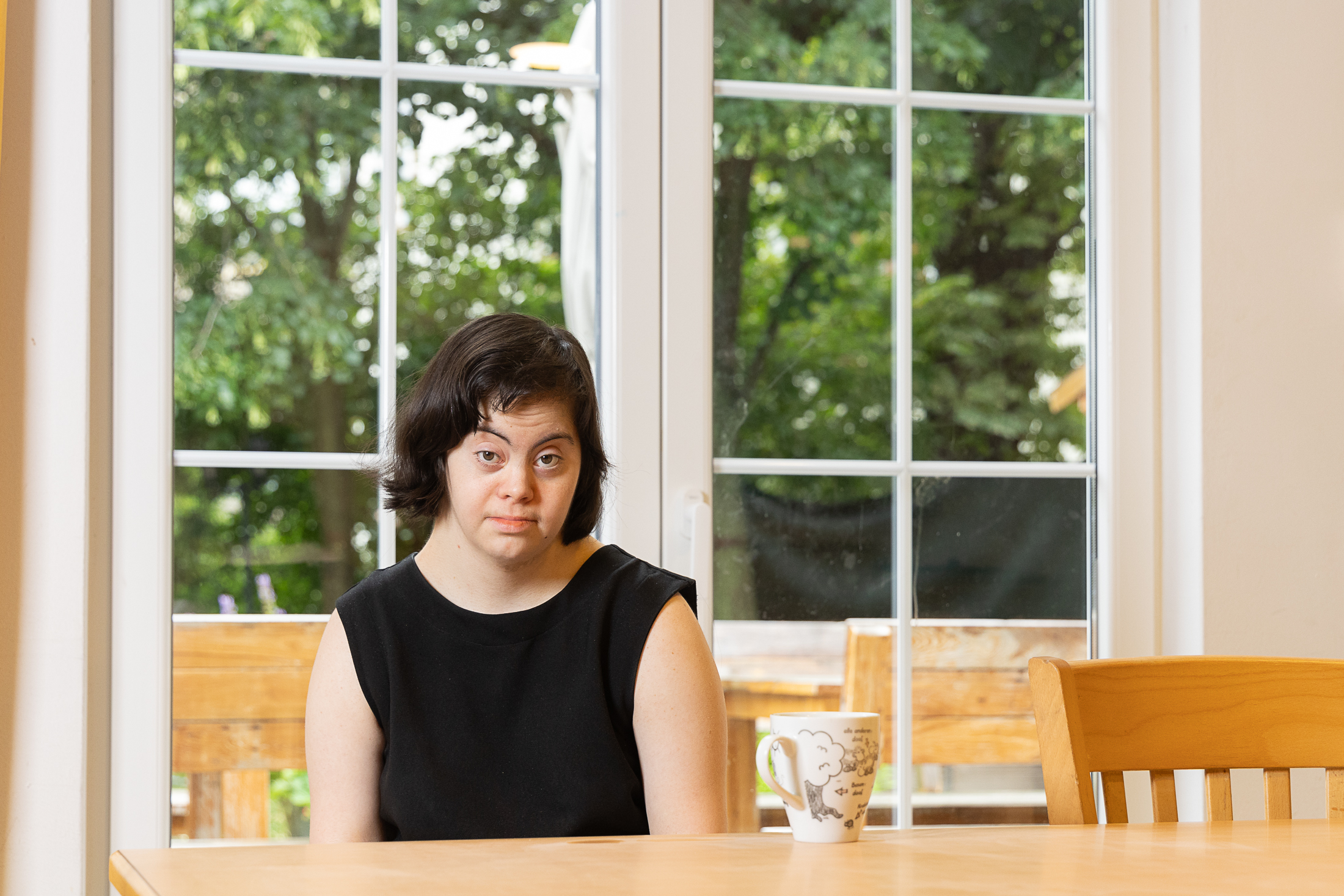 Eine junge Frau mit Downsyndrom sitzt in einem hellen Raum mit großen Fenstern an einem Tisch. Sie hat mittellange schwarze Haare und ein schwarzes aermeloses T-Shirt an. Hinter ihr vor dem Fenster sieht man einem großen Baum.