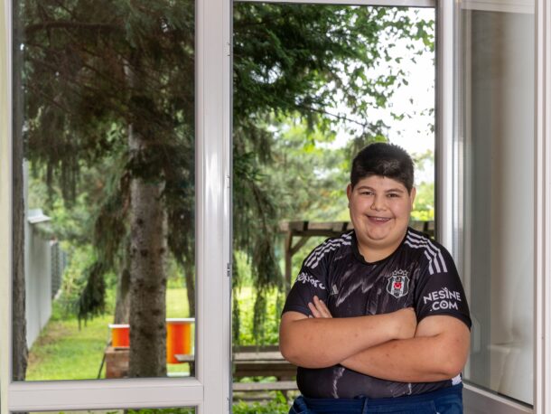 Ein junger Mann mit kurzen dunklen Haaren steht mit verschränkten Armen vor einem großen Fenster und lächelt selbstbewusst in die Kamera. Er trägt ein schwarzes T-Shirt mit weißen Aufdrucken und eine dunkle Hose. Hinter ihm ist durch das Fenster ein Garten mit Bäumen und einer Rasenfläche zu sehen. Die Atmosphäre wirkt freundlich und hell, und das natürliche Licht fällt durch das Fenster in den Raum.