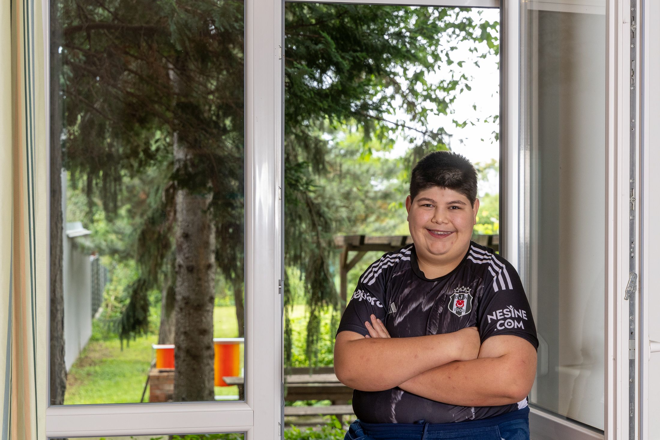 Ein junger Mann mit kurzen dunklen Haaren steht mit verschränkten Armen vor einem großen Fenster und lächelt selbstbewusst in die Kamera. Er trägt ein schwarzes T-Shirt mit weißen Aufdrucken und eine dunkle Hose. Hinter ihm ist durch das Fenster ein Garten mit Bäumen und einer Rasenfläche zu sehen. Die Atmosphäre wirkt freundlich und hell, und das natürliche Licht fällt durch das Fenster in den Raum.