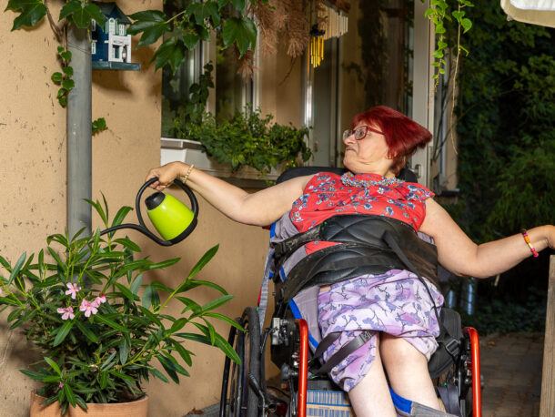 Das Bild zeigt eine Frau mit rotem Haar und bunten Oberteil und Rock. Sie sitzt in einem Rollstuhl und hat eine gruene Giesskanne in der Hand mit der sie einen bluehenden Oleanderstrauch bewaessert.