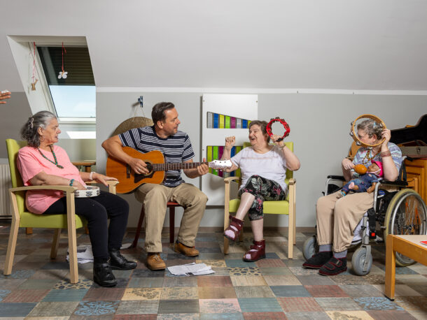 Eine Menschengruppe musiziert gemeinsam in einem freundlichen Raum. Eine Frau sitzt im Rollstuhl und spielt Tamburin, ein Mann spielt Gitarre und zwei weitere Frauen spielen auch Tamburin. Der Boden des Raums ist bunt gestaltet und es hängen noch weitere Musikinstrumente an den Wänden.