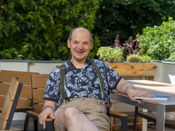 Ein lachender Mann sitzt auf Terrasse und hält ein Fotobuch in der Hand. Er trägt ein schwarz-weißes Hemd mit Pflanzen-Druck, eine beige Short und Hosenträger. Er wirkt froehlich und entspannt, im Hintergrund sieht man viele Baume.