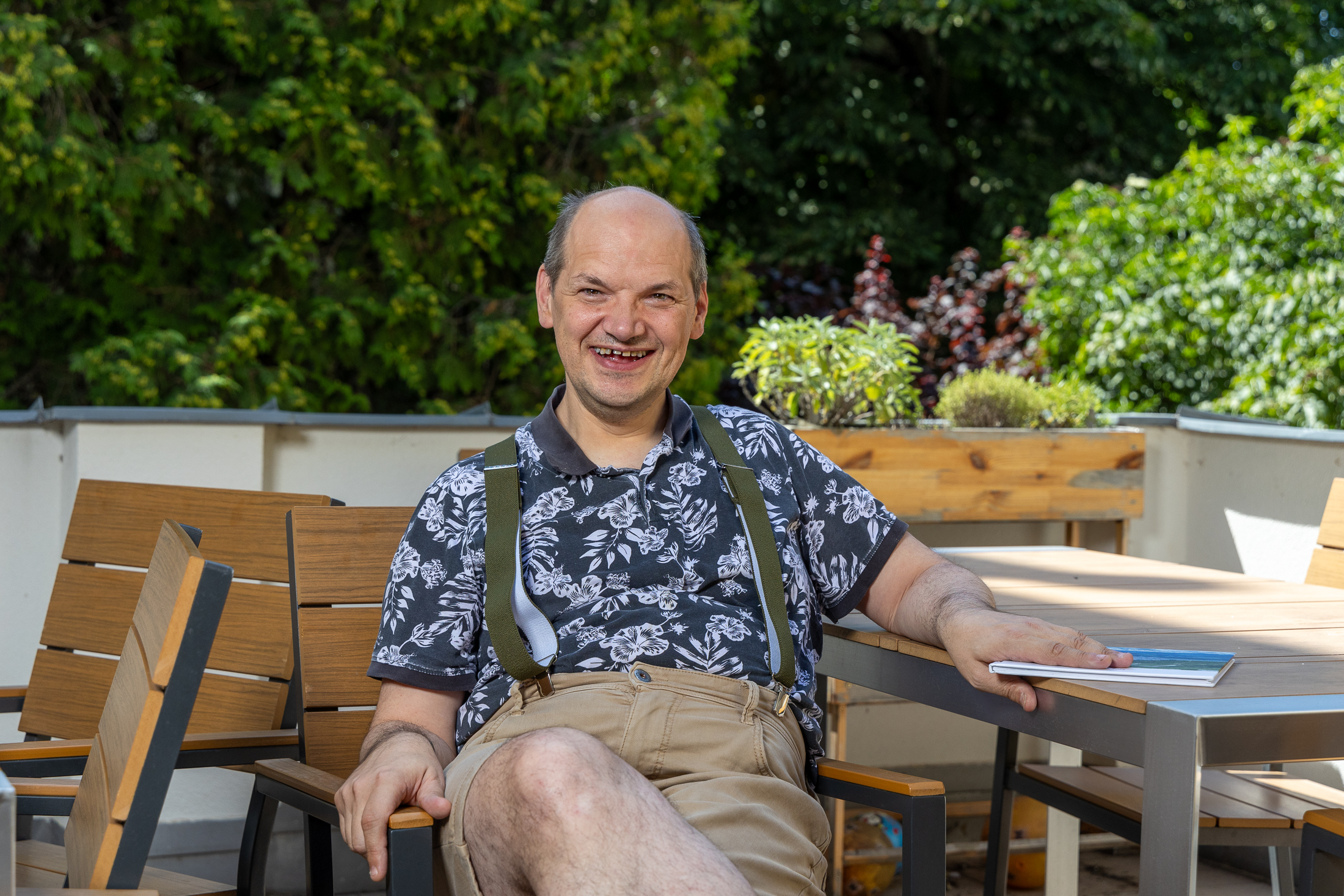 Ein lachender Mann sitzt auf Terrasse und hält ein Fotobuch in der Hand. Er trägt ein schwarz-weißes Hemd mit Pflanzen-Druck, eine beige Short und Hosenträger. Er wirkt froehlich und entspannt, im Hintergrund sieht man viele Baume.