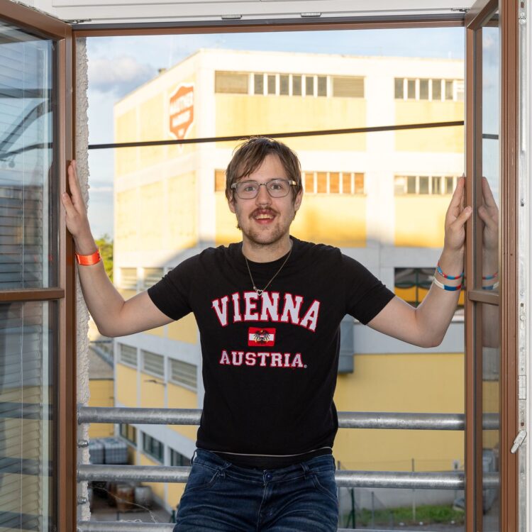 Ein junger Mann mit Brille und einem schwarzen T-Shirt lehnt sich bei geöffnetem Fenster gegen ein Geländer. Er lächelt in die Kamera und schaut sehr zufrieden aus.