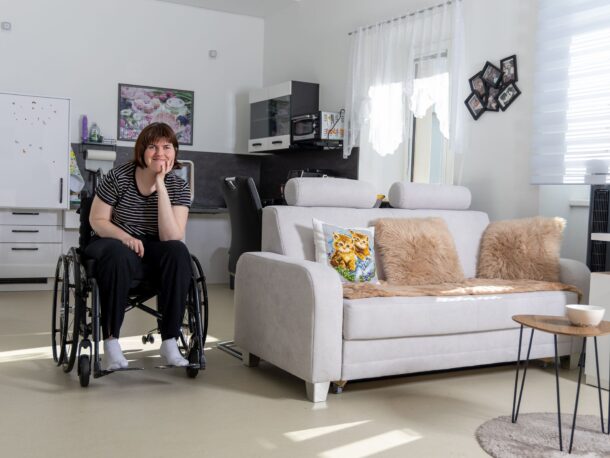 Auf dem Bild sitzt eine Frau im Rollstuhl in ihrer gemütlich eingerichteten Garconniere. Neben ihr steht eine Couch mit dekorativen Kissen, und im Hintergrund ist eine Kochnische zu sehen. Das Zimmer ist hell und freundlich gestaltet.