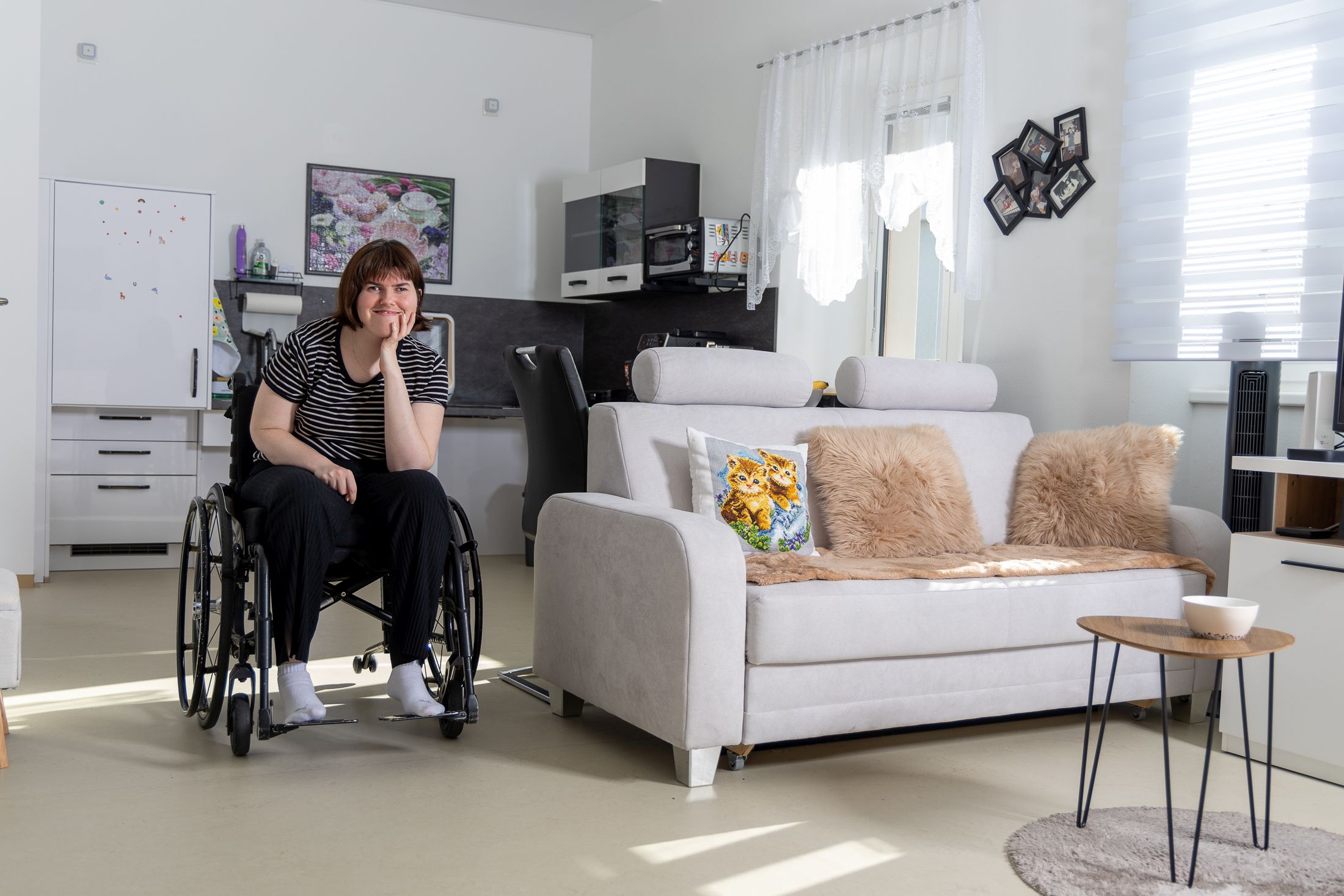 Auf dem Bild sitzt eine Frau im Rollstuhl in ihrer gemütlich eingerichteten Garconniere. Neben ihr steht eine Couch mit dekorativen Kissen, und im Hintergrund ist eine Kochnische zu sehen. Das Zimmer ist hell und freundlich gestaltet.
