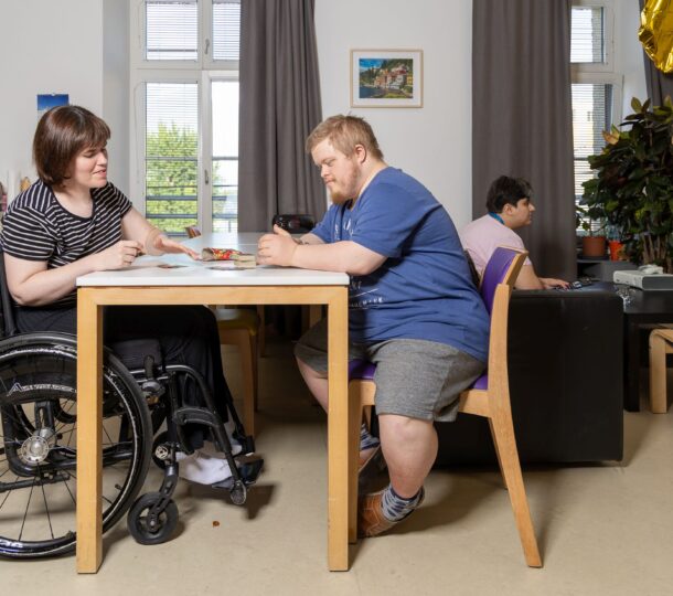 Eine Frau im Rollstuhl mit kurzen braunen Haaren und ein Mann mit blauem T-Shirt sitzen gemeinsam an einem Tisch und spielen das Kartenspiel „Uno“. Sie befinden sich im Gemeinschaftsraum. Im Hintergrund sieht eine andere Frau fern.