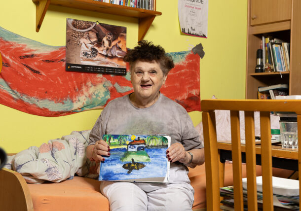 Eine lächelnde Frau sitzt auf dem Bett in ihrem Zimmer und hält ein selbstgemaltes Bild in den Händen. Auf dem Bild ist ein Haus auf einer Insel in einem See zu sehen. Im Hintergrund hängt ein Kalender und einer bemalter roter Stoff. Die Wand in ihrem Zimmer ist gelb gestrichen.