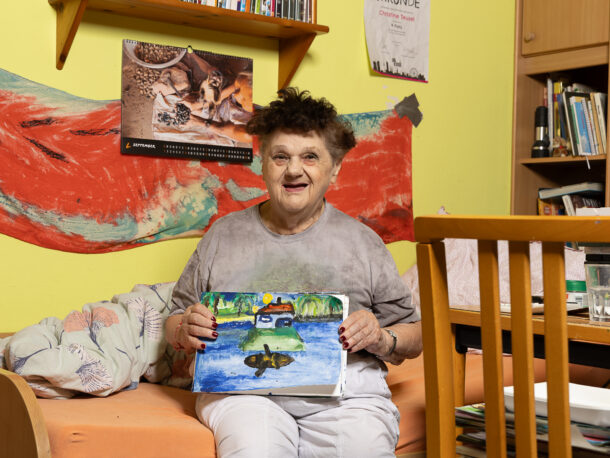Eine lächelnde Frau sitzt auf dem Bett in ihrem Zimmer und hält ein selbstgemaltes Bild in den Händen. Auf dem Bild ist ein Haus auf einer Insel in einem See zu sehen. Im Hintergrund hängt ein Kalender und einer bemalter roter Stoff. Die Wand in ihrem Zimmer ist gelb gestrichen.