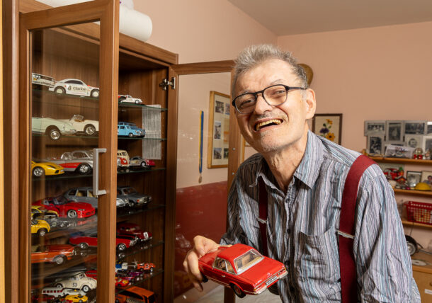 Das Bild zeigt einen Ausschnitt aus einem Zimmer. Zu sehen ist ein Kasten auf dem zwei Urkunden, zwei Medaillen, Pokale und weitere Gegenstände stehen. Die Wand ist mit Bildern von Pflanzen dekoriert und aus dem Fenster blickt man ins Grüne.