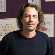 Ein Mann mit längeren dunkeln Haaren steht vor einer violetten Wand mit vielen Bildern. Er traegt einen schwarzen Sweater und Jeans.
